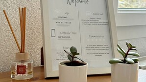 A framed welcome sign with check-out instructions, a small potted plant, and a reed diffuser on a wooden surface. - Studio Cosy (Mende)