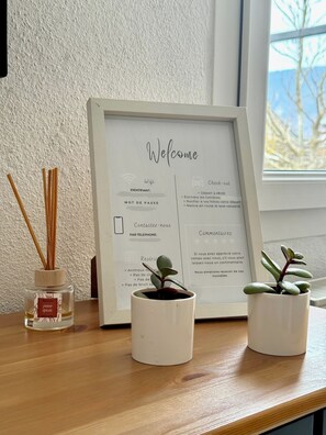 A framed welcome sign with check-out instructions, a small potted plant, and a reed diffuser on a wooden surface. - Studio Cosy (Mende)