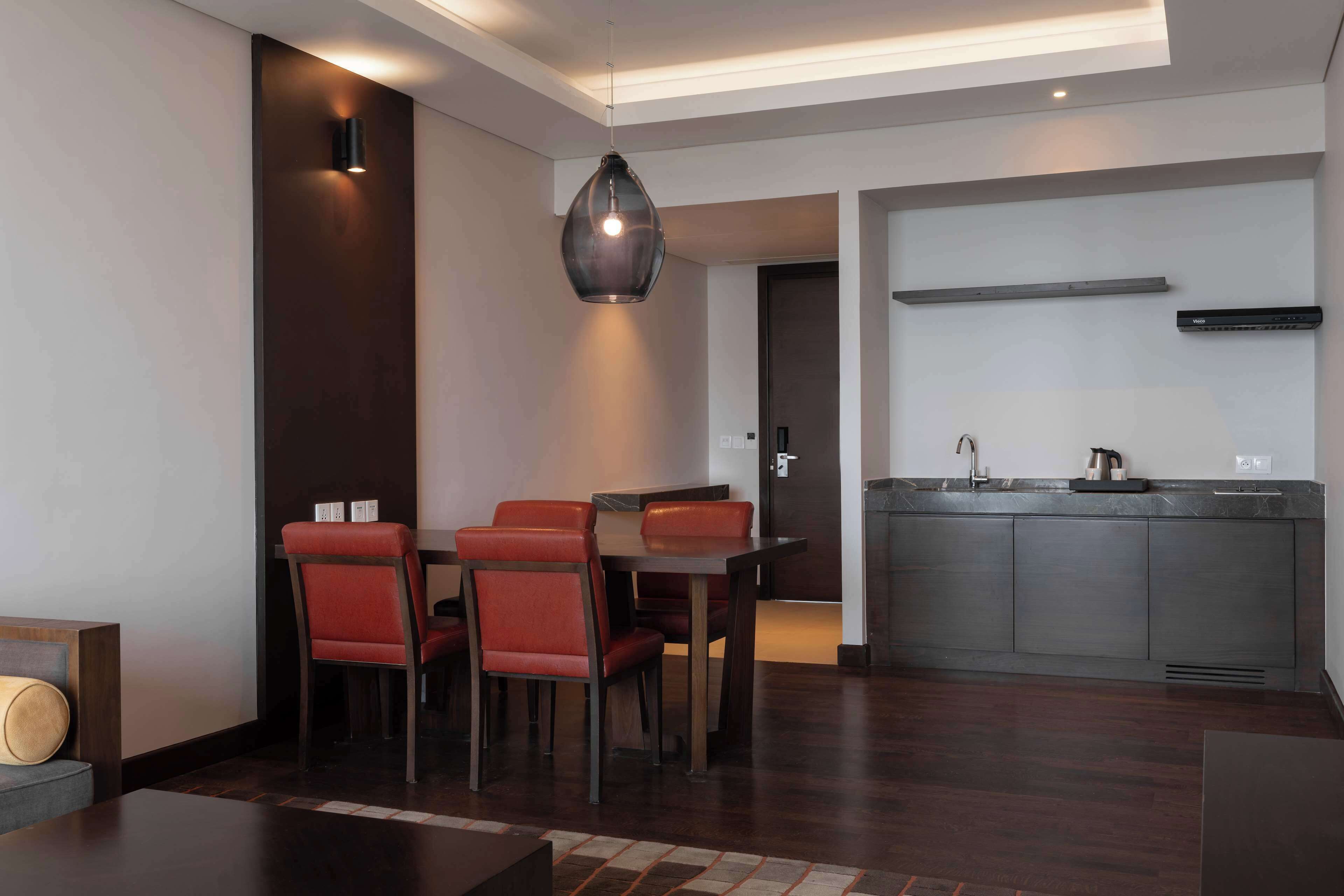 A modern dining area with a dark wood table and red chairs, a pendant light, and a kitchen with a dark countertop and stainless steel appliances.