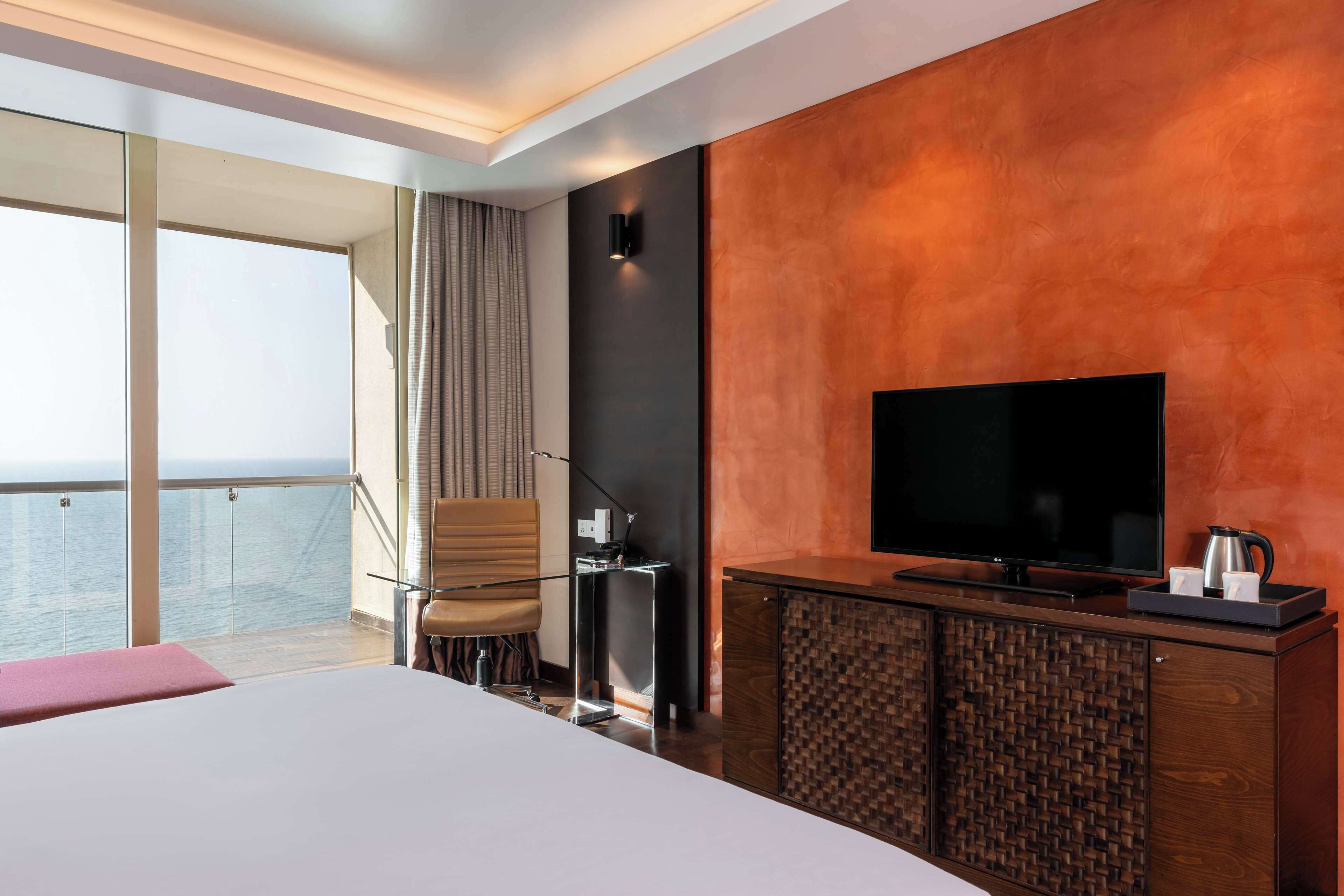 A modern hotel room with a large bed, a flat-screen TV, a wooden cabinet, and a view of the sea through large windows.