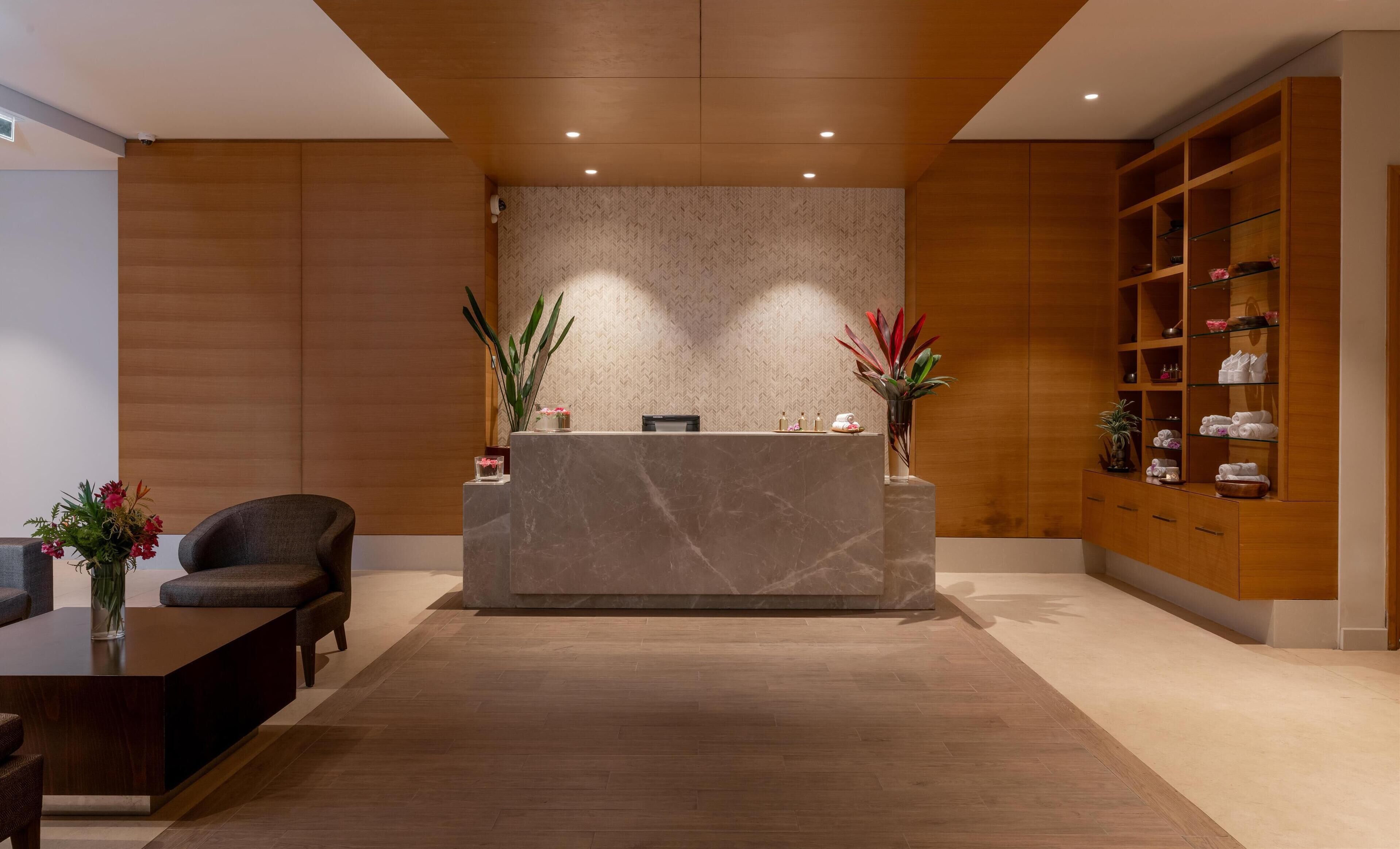 A reception area with a marble counter, two potted plants, a sofa, and a wooden shelving unit with towels and toiletries.