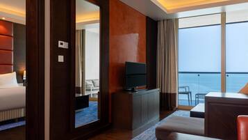 Una habitación de hotel moderna con una cama grande, un televisor sobre un mueble de madera, una zona de estar con vista al mar y un balcón con mesa y sillas.
