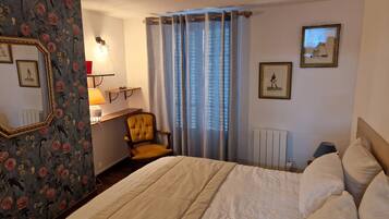 A hotel room with a bed, a chair, a desk with a lamp, a radiator, and curtains.