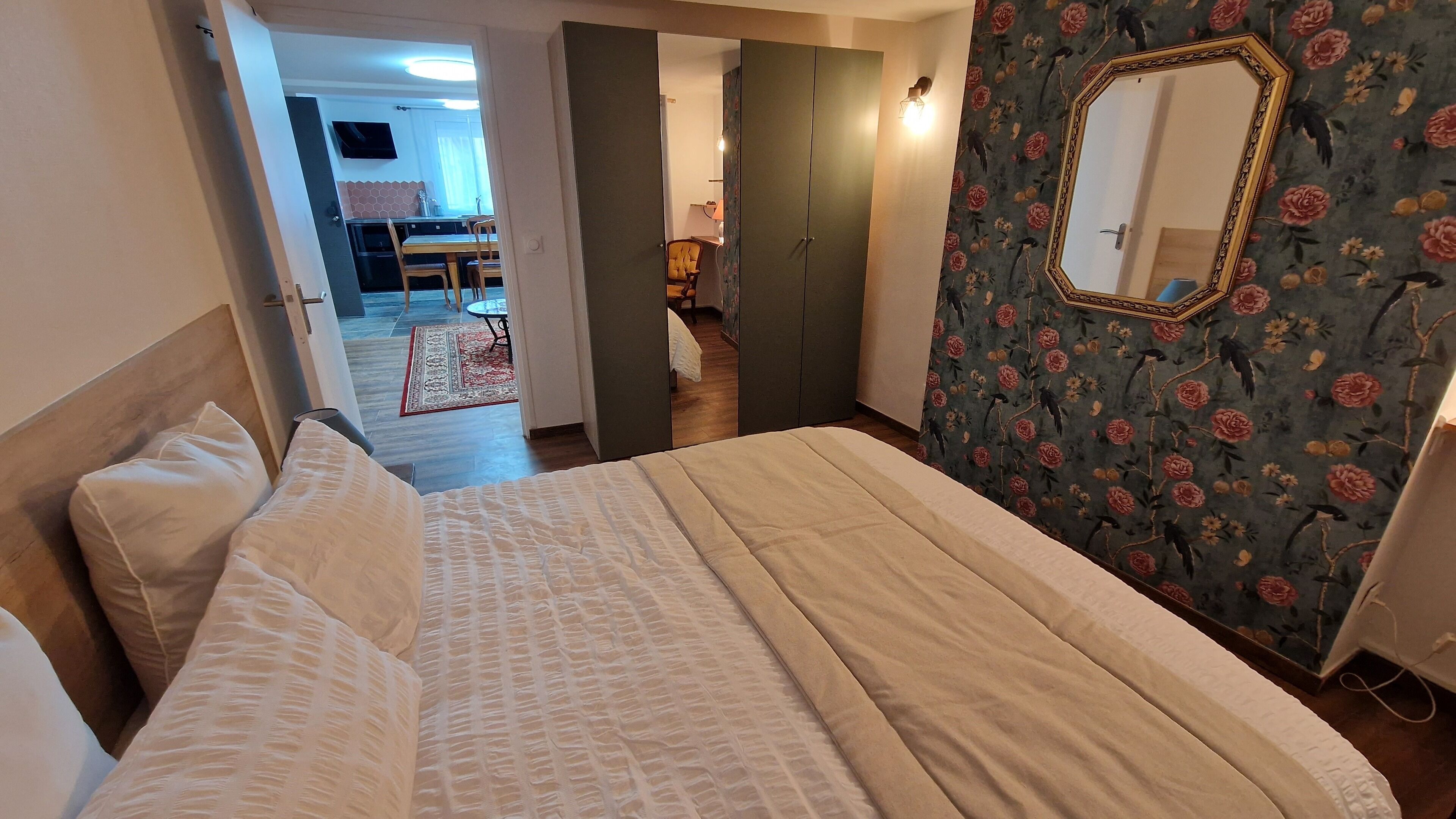 A bedroom with a bed, a wardrobe, and a mirror. There is a dining area visible through an open door.