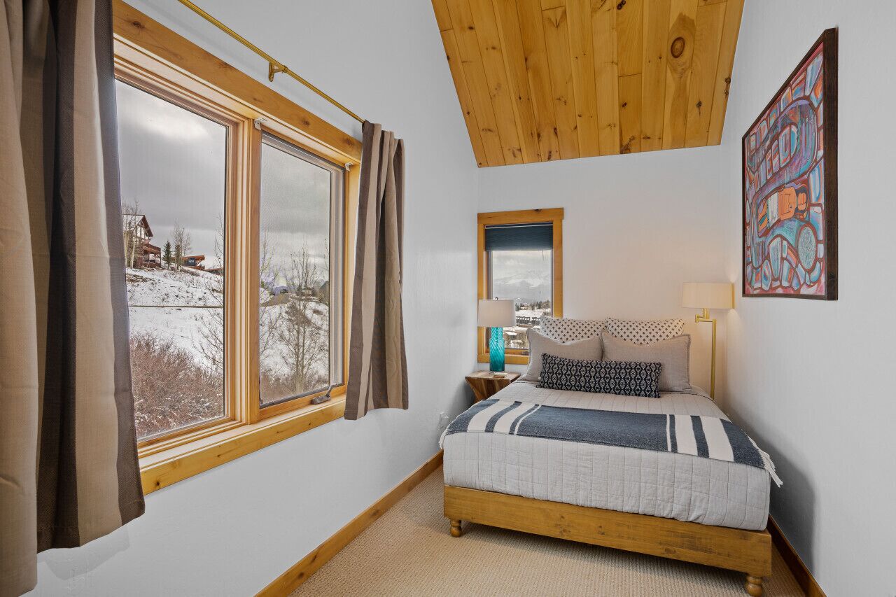 A bedroom with a wooden ceiling, a large window with curtains, a bed with patterned bedding, a bedside table with a lamp, and a painting on the wall.