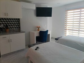 Habitación de hotel con kitchenette, televisor montado en la pared, cama y ventana con persianas.