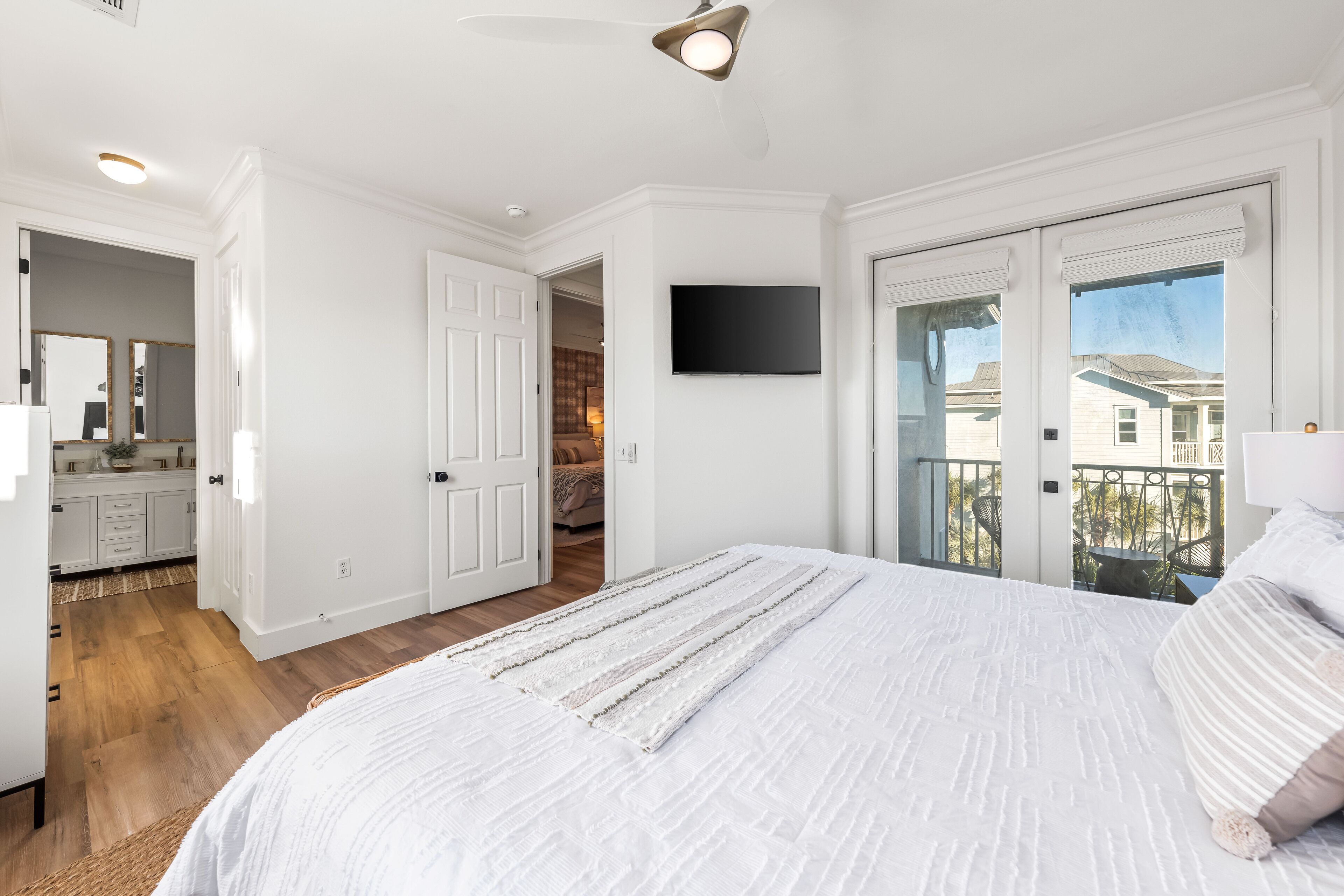 A bedroom with a large bed, a TV mounted on the wall, and a balcony with a view of houses.