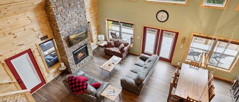 A spacious living area with a stone fireplace, wooden floors, and large windows offering a view of the outdoors.