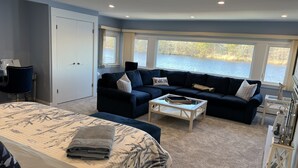 A modern living room with a large bed, a dark blue sofa, a coffee table, and a view of a lake through large windows. - Waterfront home, deck, bbq, fire pit, boats, hot tub, and more!  (Oakdale)