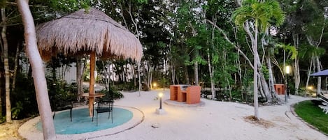 A thatched-roof pavilion with seating, surrounded by trees and a sandy area with a barbecue setup.