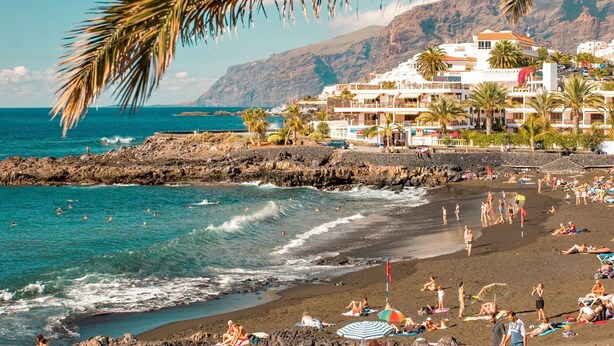 A beach with a rocky shoreline, people sunbathing, and a building with balconies overlooking the sea.