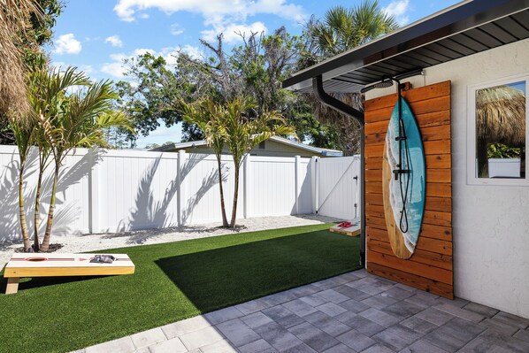 A backyard with artificial turf, a wooden bench, a surfboard, and a white fence.
