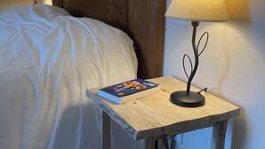 Een houten nachtkastje met een lamp, een boek en een bed met wit beddengoed.