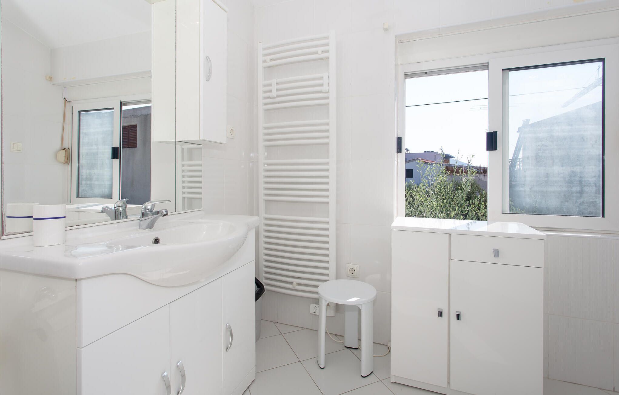Ein modernes Badezimmer mit weißen Armaturen, einem großen Spiegel, einem Fenster mit Blick auf Gebäude und einer Heizung.