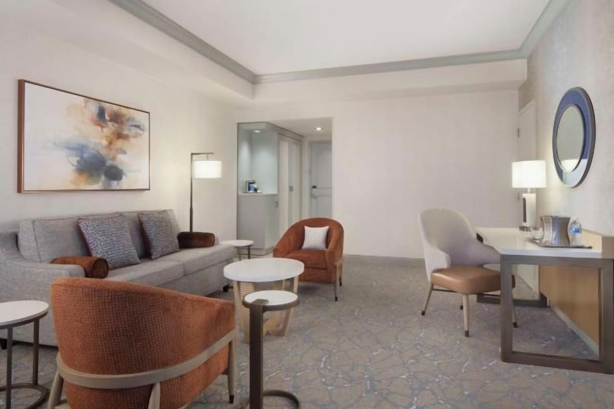 A modern hotel room with a grey sofa, orange armchair, a small round table, and a desk with a lamp.