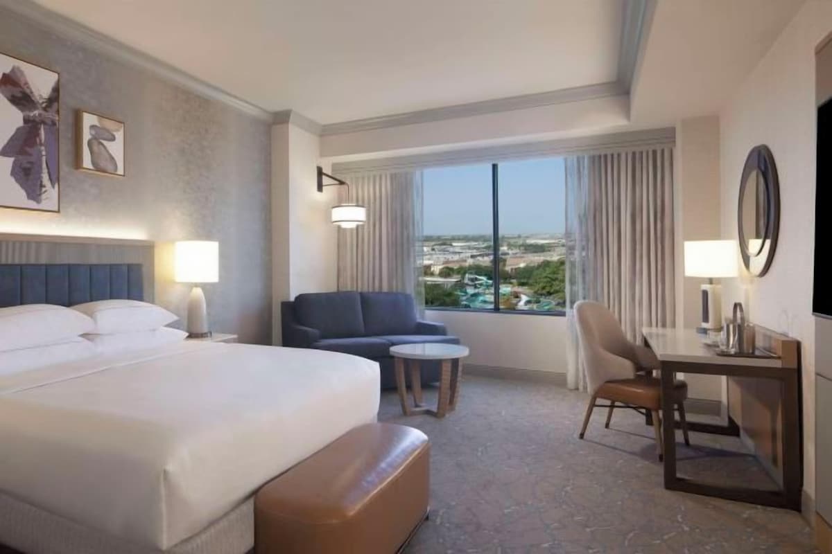 A hotel room with a bed, a desk, a chair, a small table, and a view of the city.