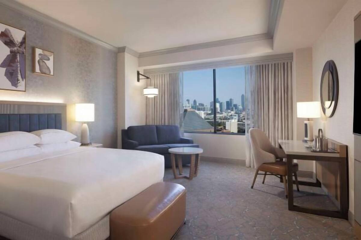 Une chambre d’hôtel avec un lit, un canapé, une petite table, une chaise, un bureau et une grande fenêtre donnant sur la ville.