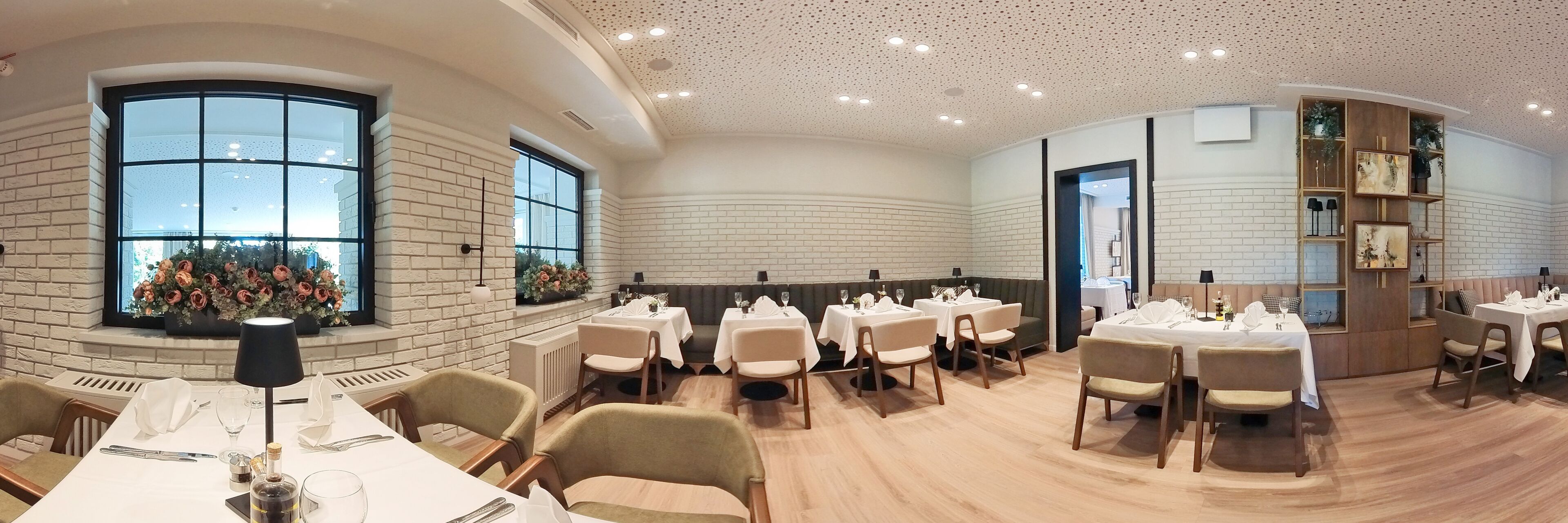 A restaurant with white brick walls, large windows, and a modern ceiling design. Tables are set with white tablecloths and chairs.