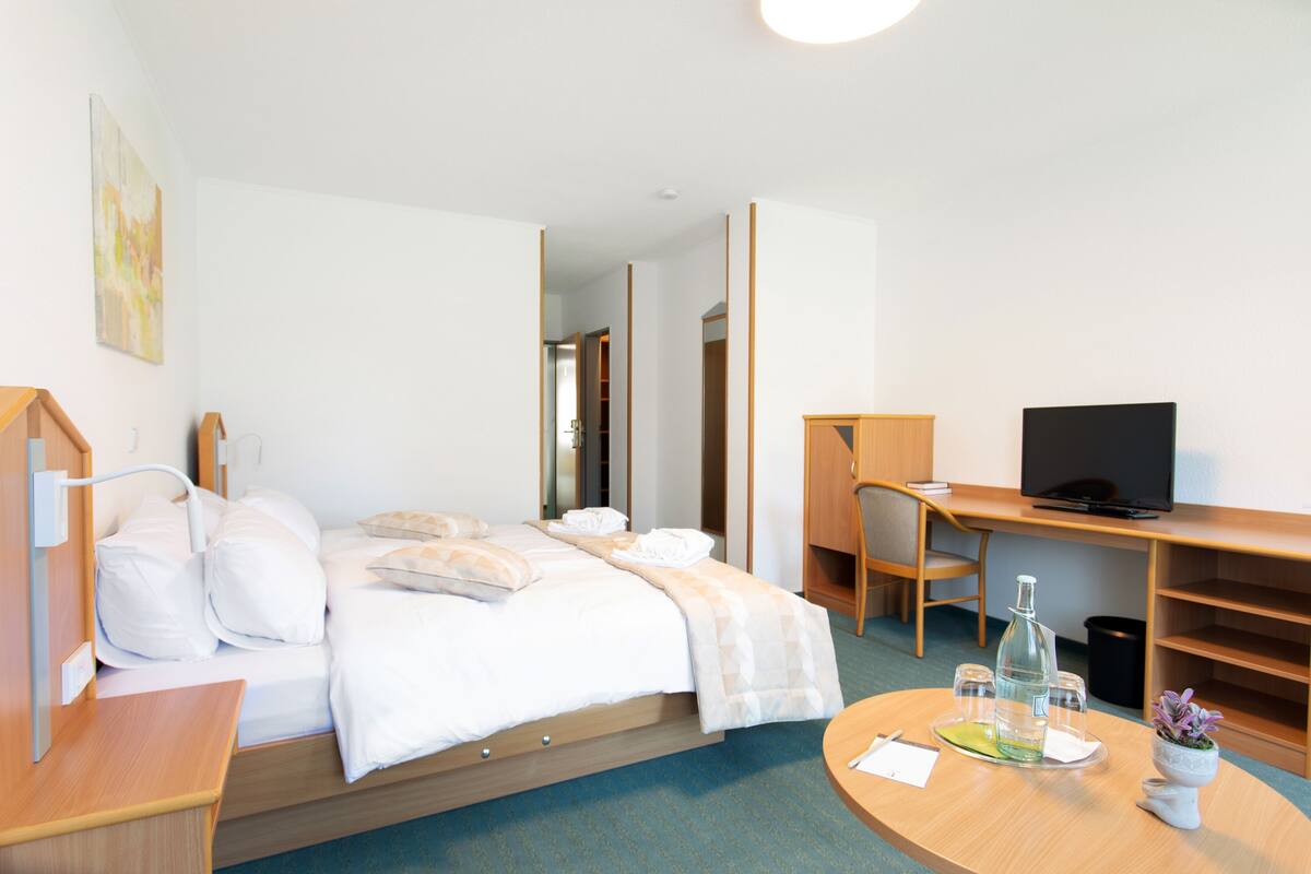 A hotel room with a bed, a desk, a television, and a small table with a bottle and glasses.