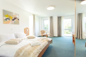 A hotel room with a large bed, a desk, and a chair. There are windows with curtains, a painting on the wall, and a carpeted floor. - Hotel Bessunger Forst (Rossdorf)