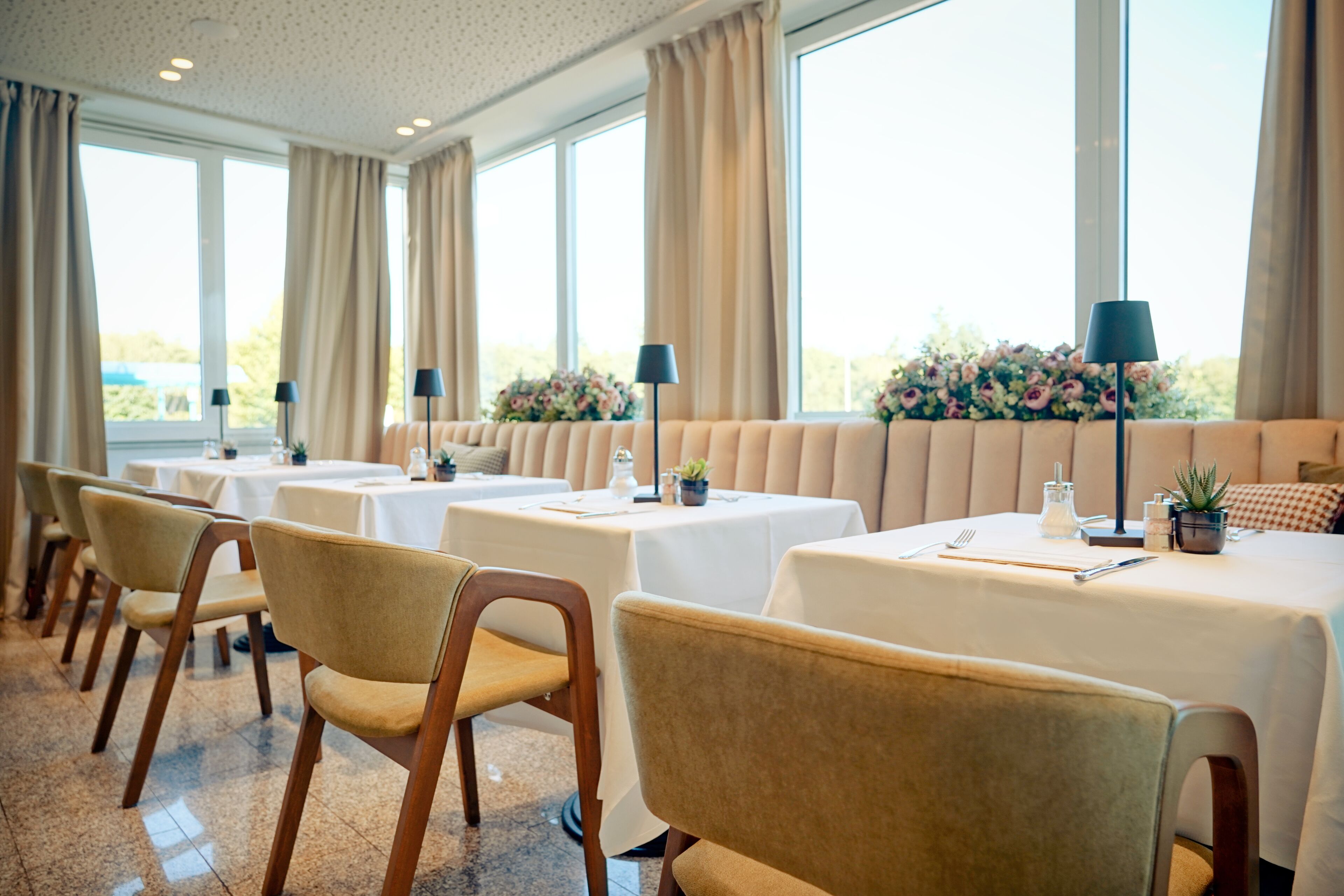 Een eetruimte met tafels en stoelen, grote ramen met gordijnen en bloemarrangementen op de vensterbanken.