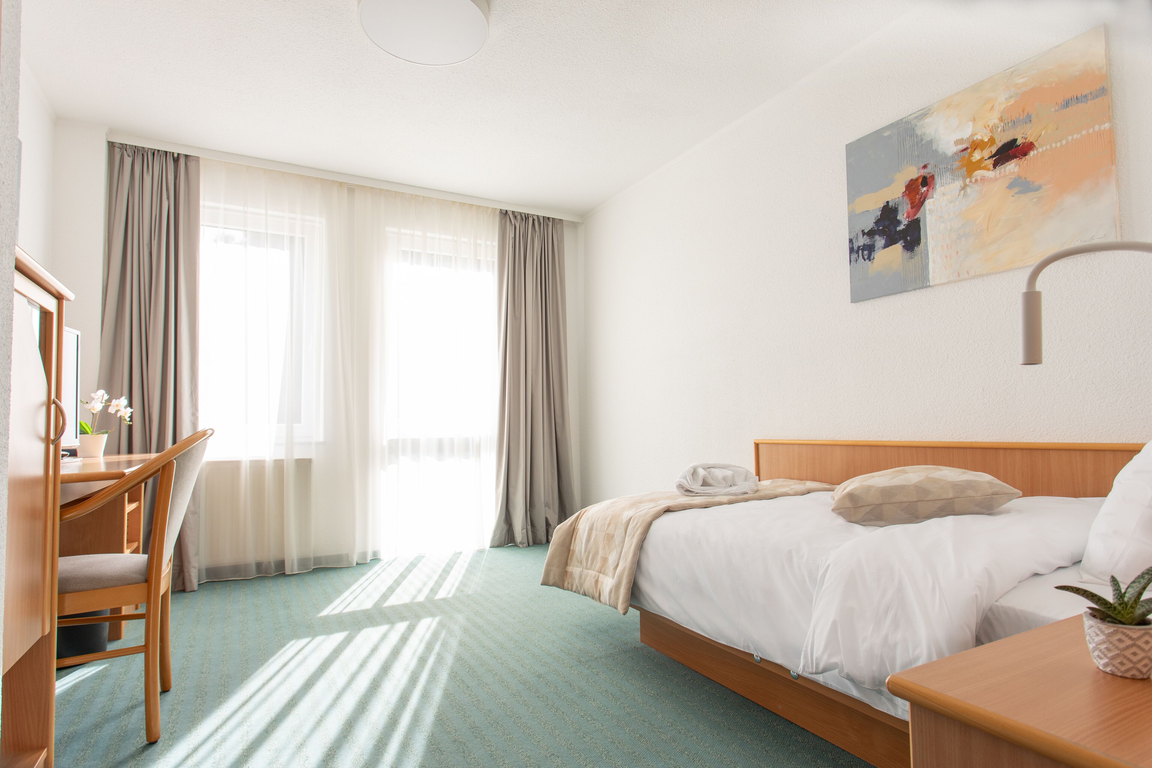 Habitación de hotel con cama, escritorio, silla, mesita de noche con una planta y un cuadro en la pared.