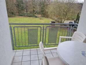 A balcony with a table and chairs, a metal railing, and a view of a grassy yard with trees and a wooden structure. - Hotel Bessunger Forst (Rossdorf)