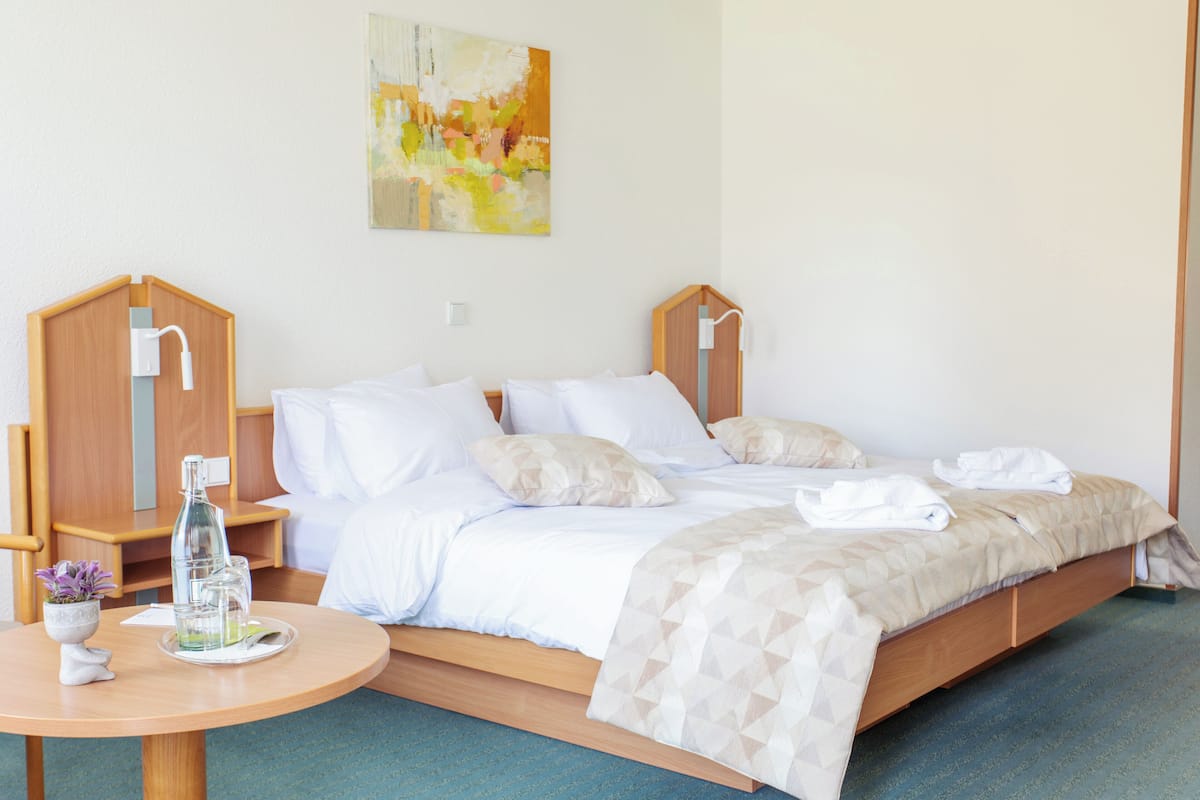 A neatly made bed with white linens, a bedside table with a glass of water, and a painting on the wall.