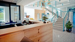 A modern reception area with a wooden desk, a staircase with teal railings, and a view of the city. - Hotel Bessunger Forst (Rossdorf)