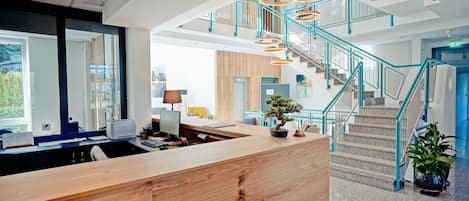 A modern reception area with a wooden desk, a staircase with teal railings, and a view of the city.