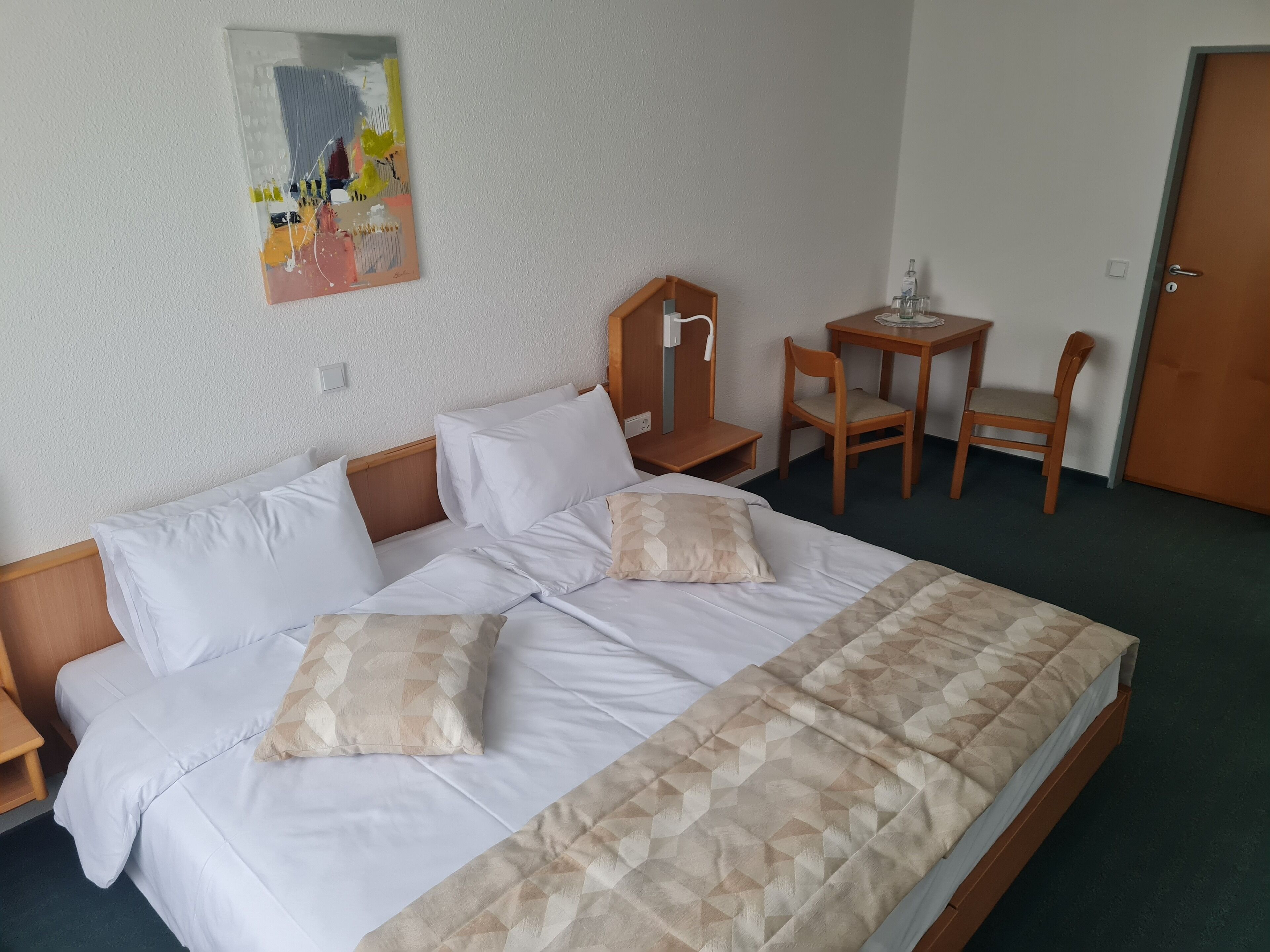 Un lit bien fait, avec du linge de lit blanc et deux coussins décoratifs, une petite table en bois avec une chaise, et un tableau accroché au mur.