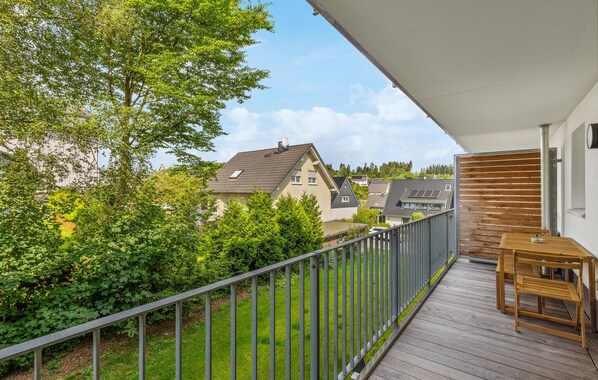 Ein Balkon mit Holztisch und -stühlen, einem Metallgeländer und Blick auf benachbarte Häuser und Bäume.