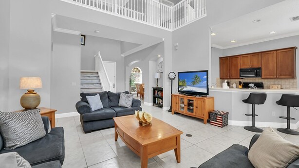 A spacious living area with a sofa, coffee table, and a TV. A kitchen with wooden cabinets and a bar area is visible. - Mediterranean Style Family Villa Close To Disney (Kissimmee)