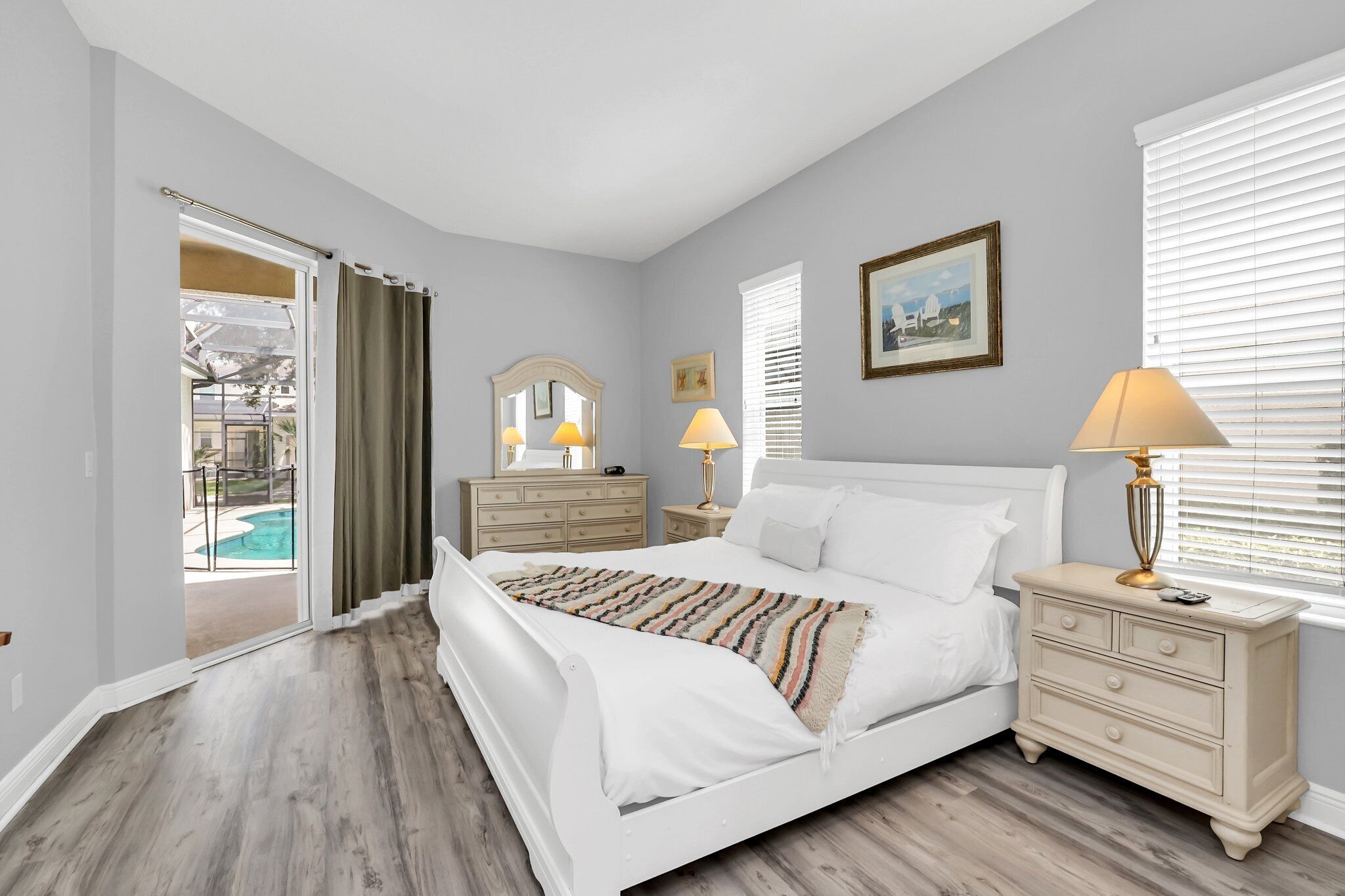 A bedroom with a bed, bedside tables, a dresser, a window with blinds, and a door leading to a pool area.