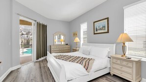 A bedroom with a bed, bedside tables, a dresser, a window with blinds, and a door leading to a pool area. - Mediterranean Style Family Villa Close To Disney (Kissimmee)