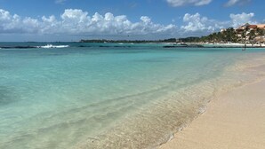 Una spiaggia con acque cristalline color turchese, un litorale sabbioso e una vista lontana di un resort con palme e edifici.