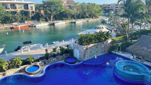 Area piscina di un resort con varie attrazioni acquatiche, un molo con barche e vista sugli edifici e sul verde circostante.