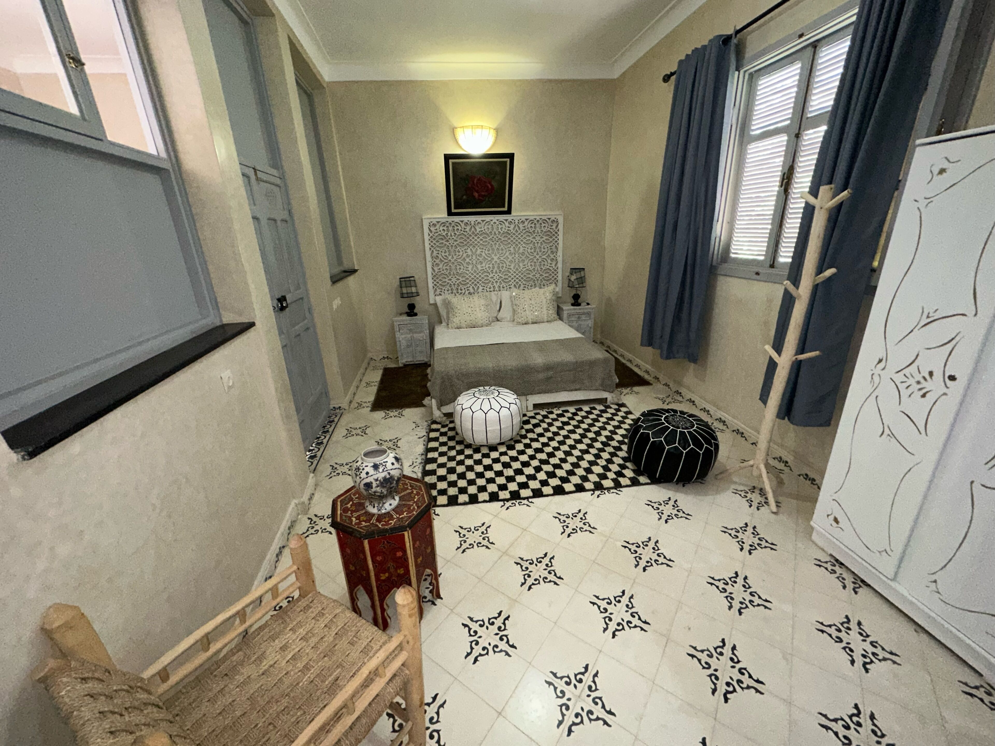 A bedroom with a checkered floor, a bed with a patterned headboard, a nightstand, a small table with a vase, and a chair.