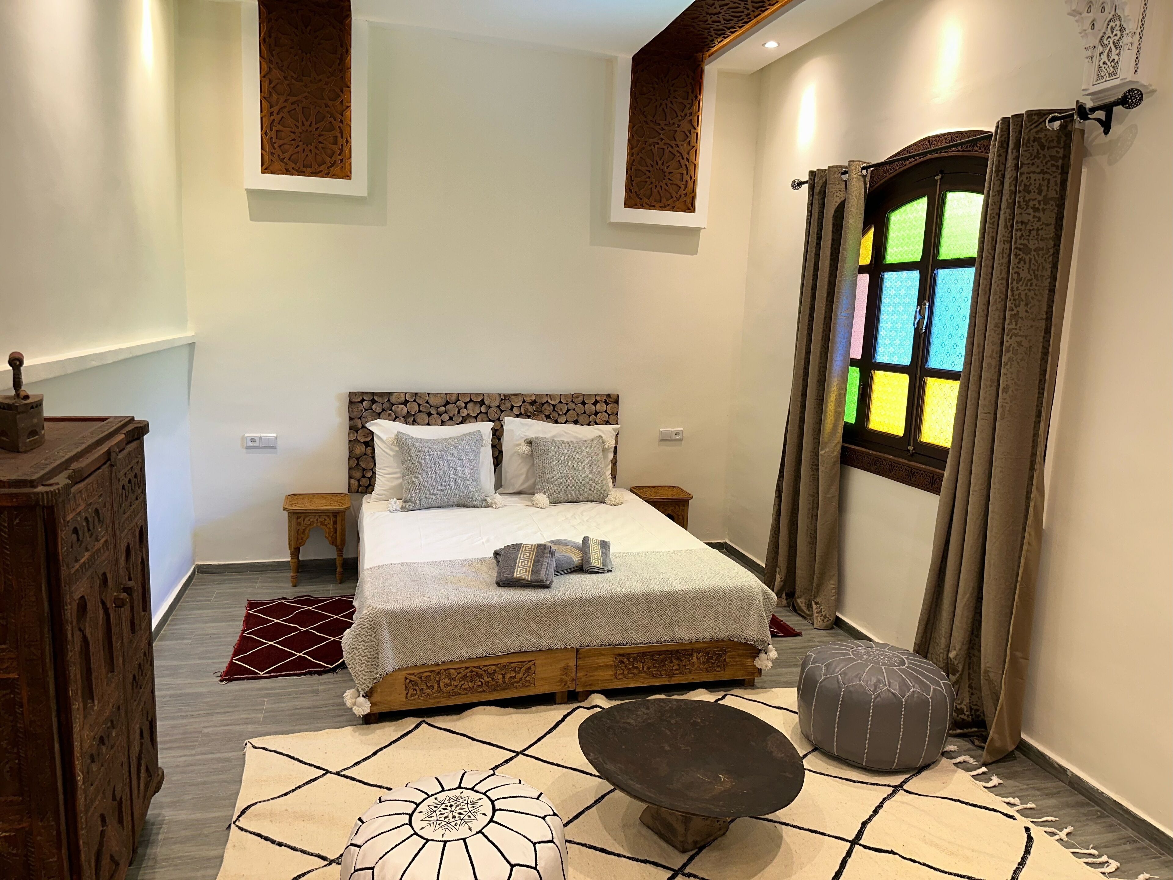 A bedroom with a bed, a wooden headboard, a nightstand, a small table, a decorative rug, a bench, and a window with curtains.
