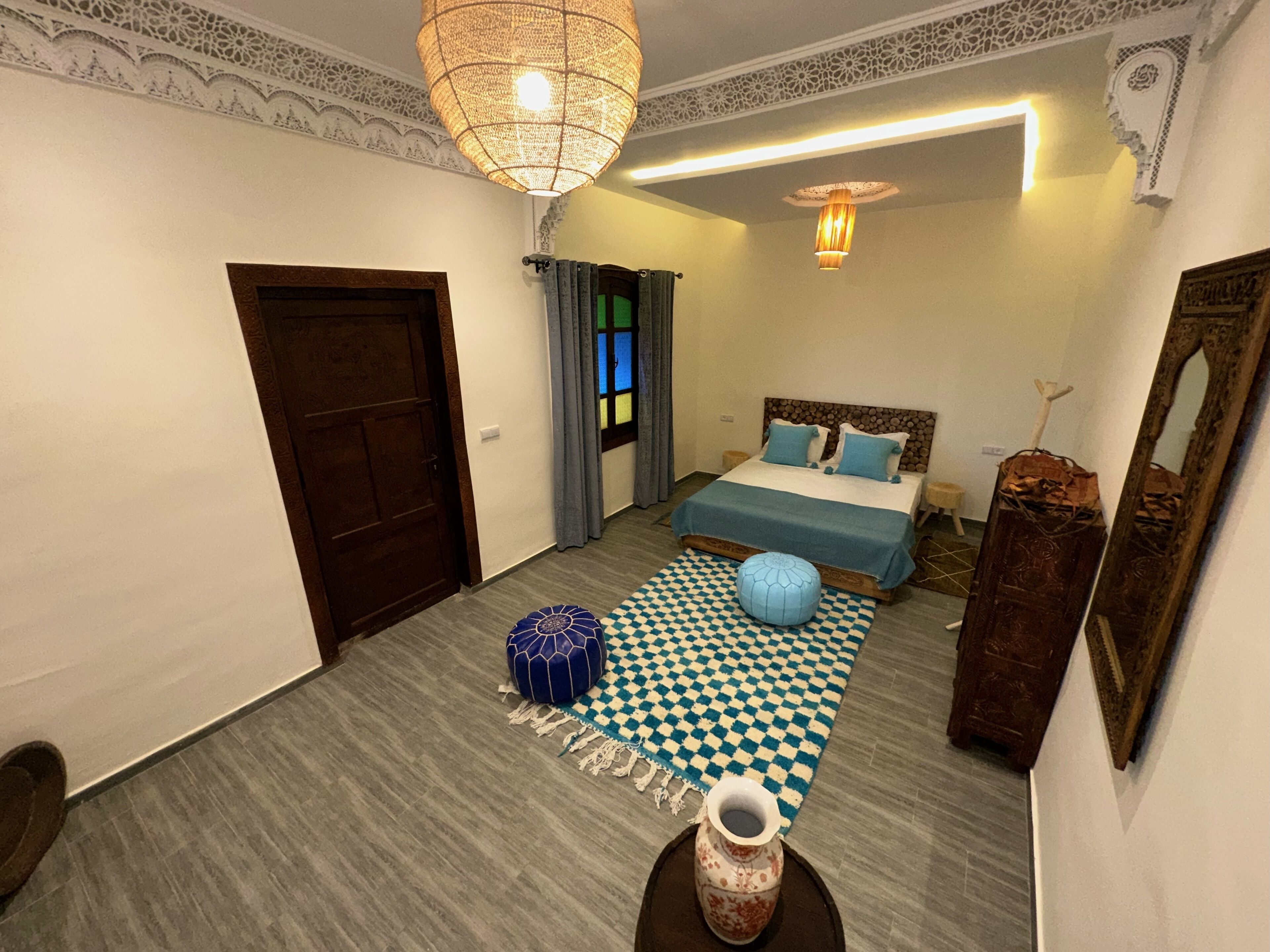 A bedroom with a checkered rug, a blue ottoman, a vase, and a bed with blue bedding.