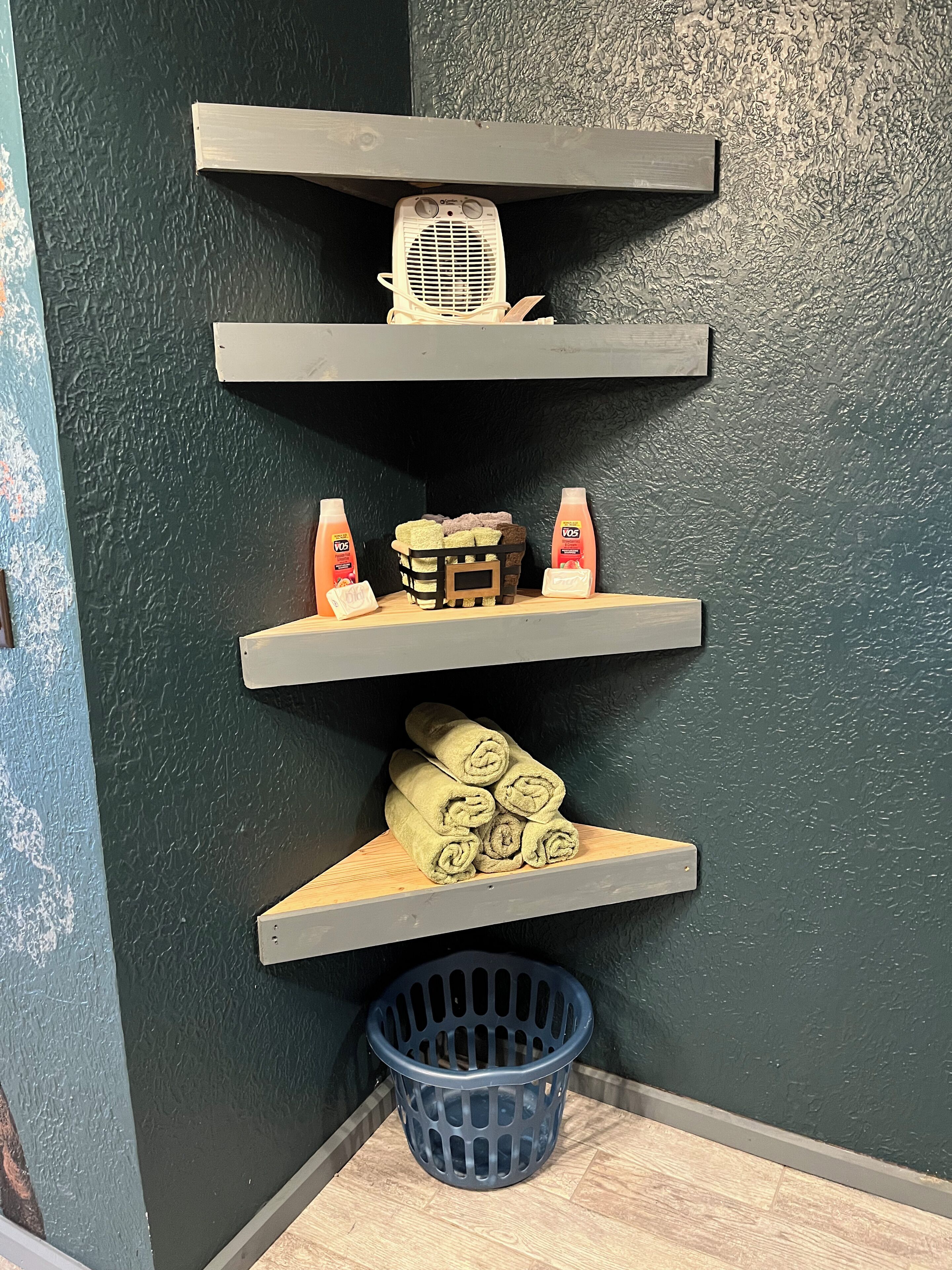 Un rincón con tres estantes de madera contra una pared texturizada, que sostiene una radio antigua, artículos de aseo y toallas enrolladas.