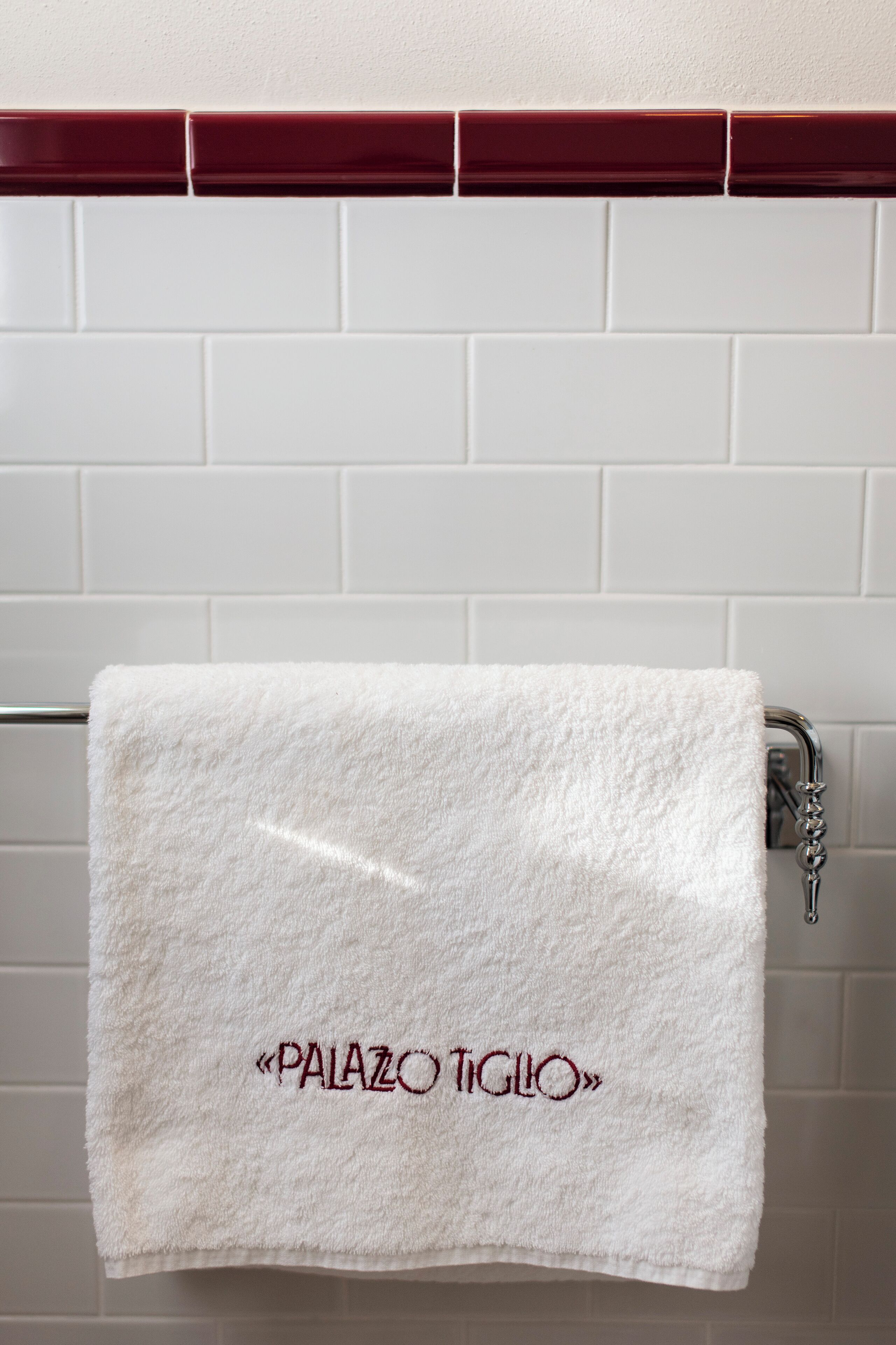 A white towel with "PALAZZO TIGIO" embroidered on it hanging on a metal towel rack against a white tiled wall.