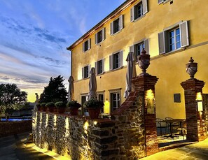 Ein zweistöckiges Gebäude mit einer Steinwand und mehreren Fenstern mit Fensterläden. Vor der Fassade befindet sich Sitzgelegenheiten mit Sonnenschirmen und im Hintergrund ein kleiner Pool.