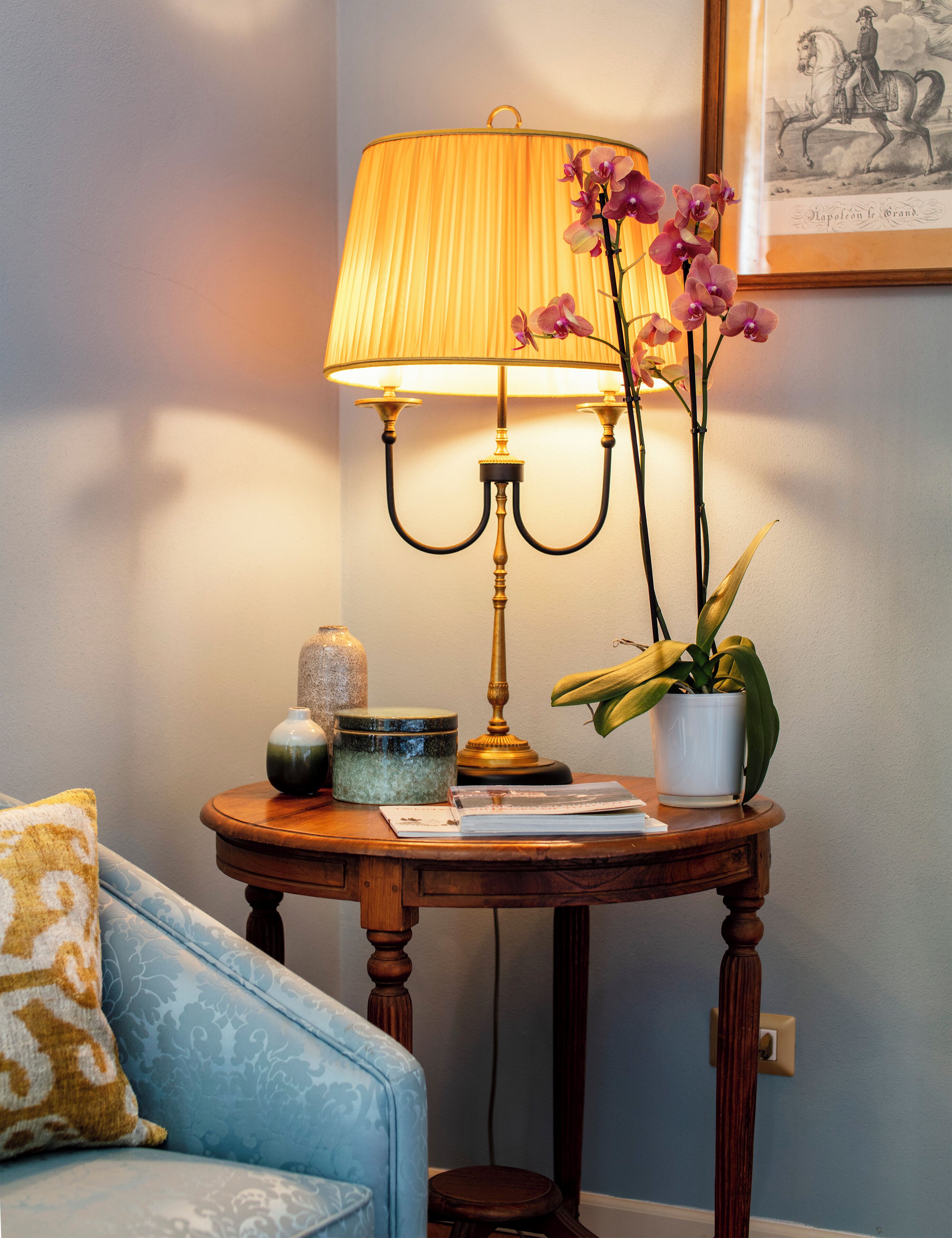 Une table ronde en bois, sur laquelle se trouvent une lampe en laiton, une orchidée en pot, un petit vase et un tableau encadré accroché au mur.