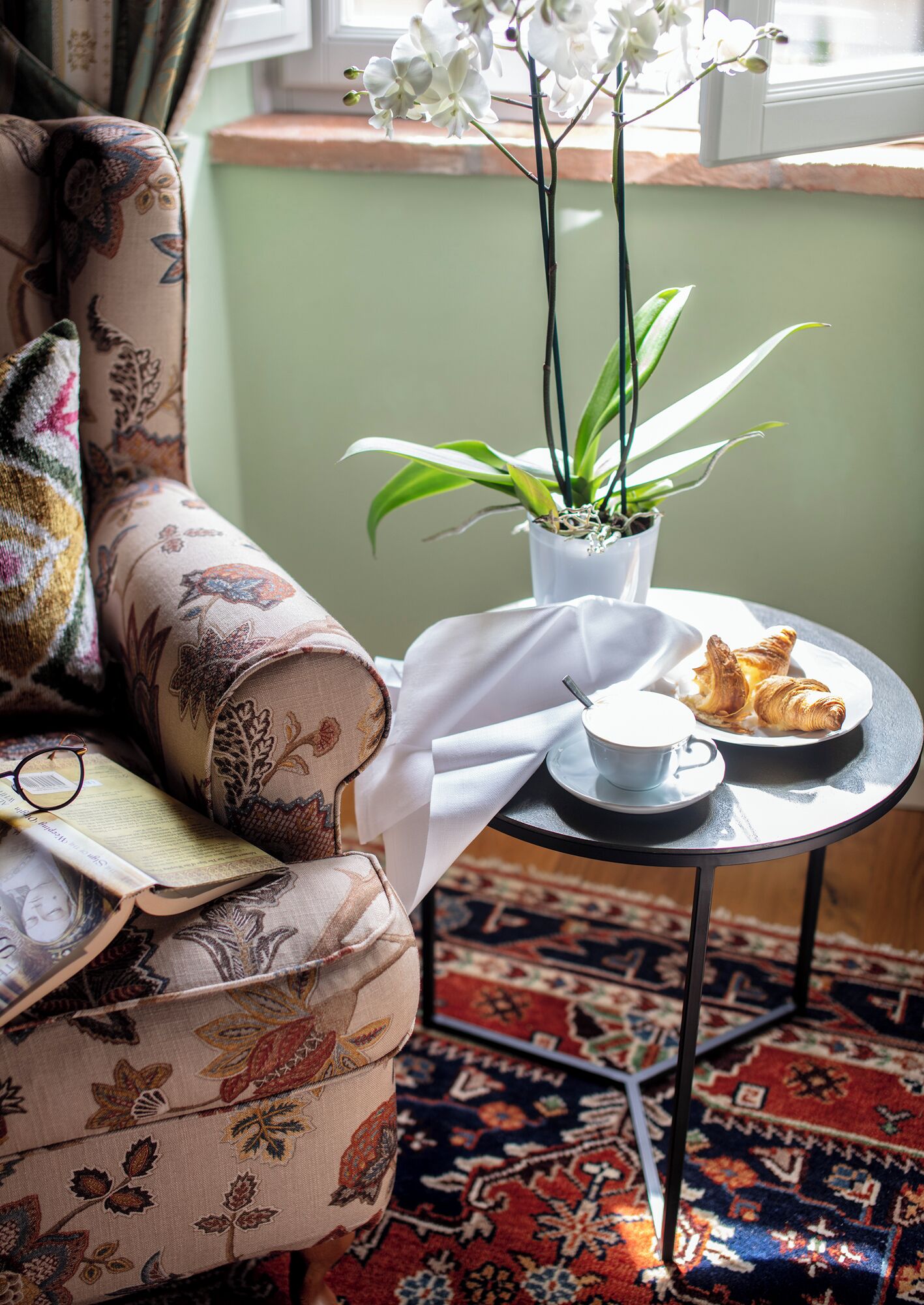 A cozy room with a patterned sofa, a small round table with a cup and pastries, and a potted plant with white flowers.
