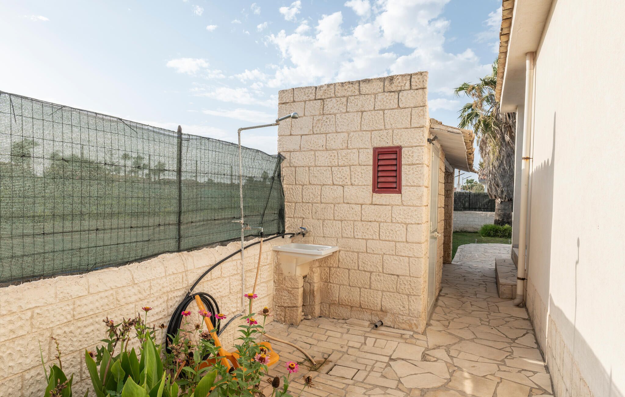 A small outdoor area with a stone wall, a metal fence, a white sink, and a garden with flowers.