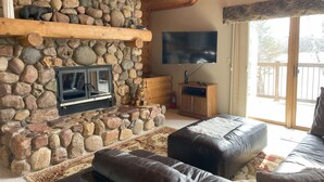Una chimenea de piedra con un televisor encima, un banco de madera y un sofá en una sala de estar acogedora con un ventanal grande.