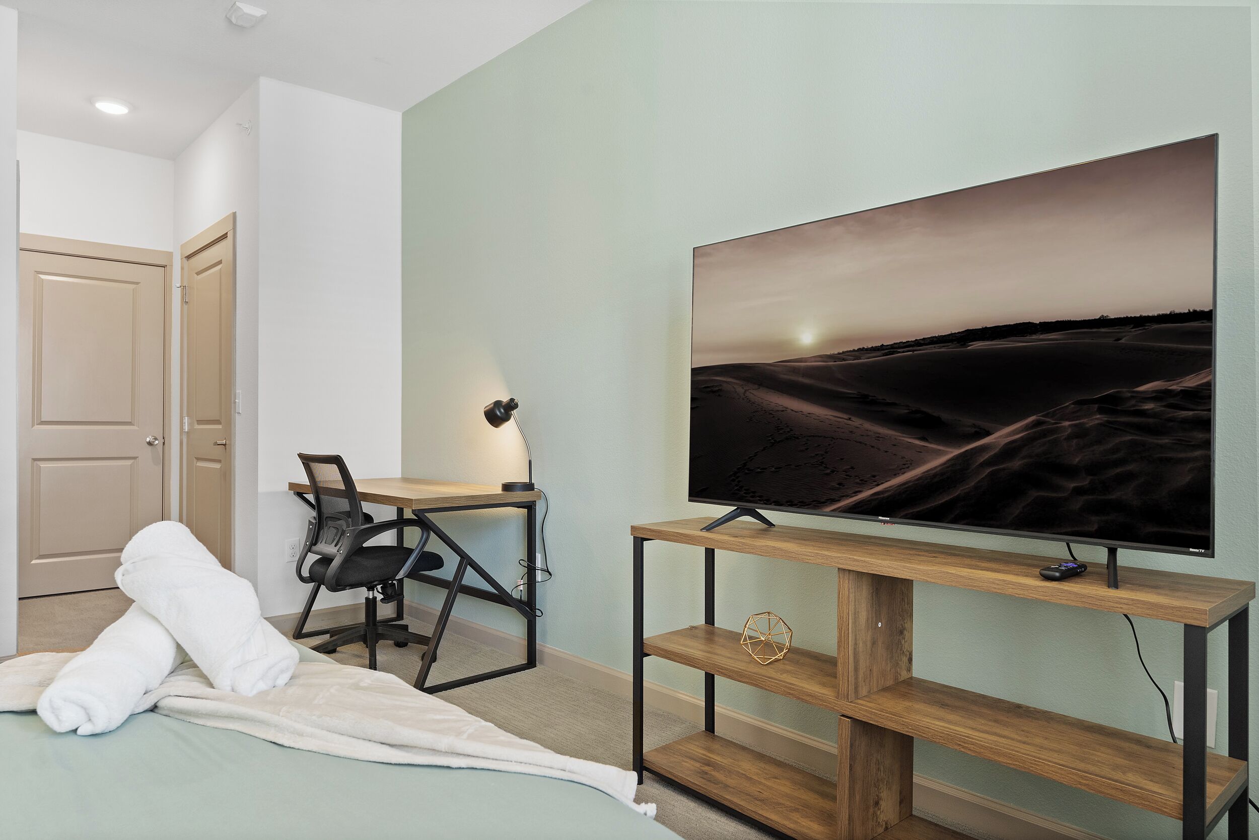 Une chambre d’hôtel avec un lit, un bureau avec une chaise, un téléviseur posé sur un meuble en bois et une porte.