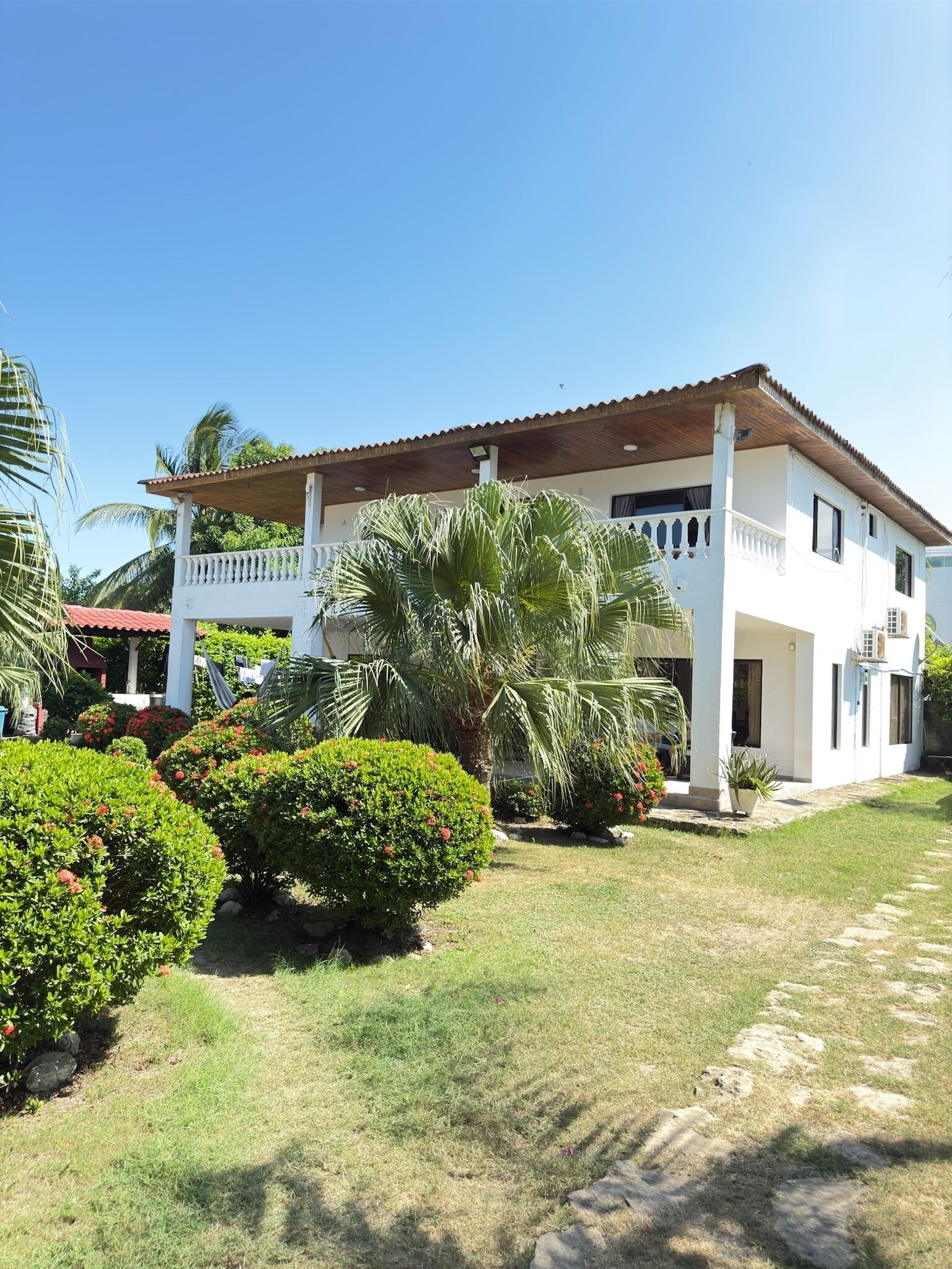 A white two-story house with a red-tiled roof, surrounded by well-maintained lawns and palm trees.