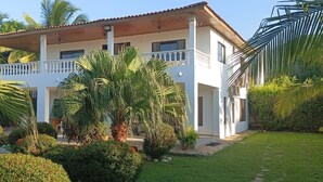 Una casa de dos pisos con balcón, rodeada de exuberante follaje y palmeras.